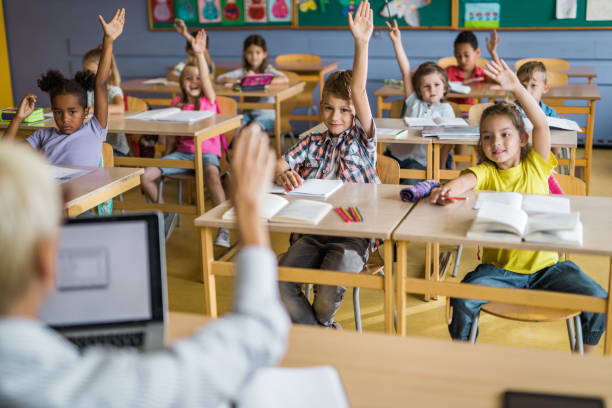 istock Large group of elementary students raising arms to answer the teacher's question on a class. 1005285462