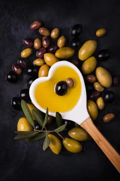 ladle shaped like a heart full of oil and some types of olives on the dark background
