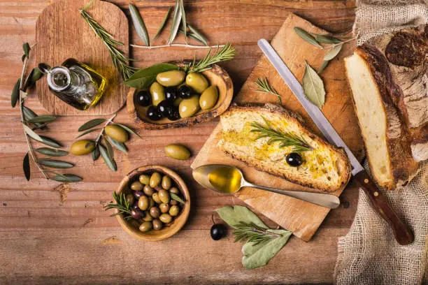 on the rustic wooden table, a spoon full of oil some olives and sliced bread