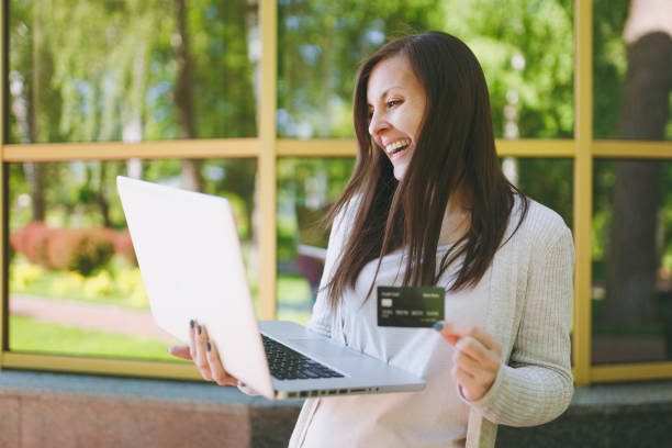 young girl in light casual clothes holding credit card. woman working on modern laptop computer near mirror building with tree reflection in street outdoors. mobile office. freelance business concept. - child office chaos computer monitor imagens e fotografias de stock