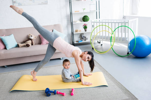 istock young mother practicing yoga in One Legged Downward-Facing Dog pose while her little child sitting on mat at home 1005335076