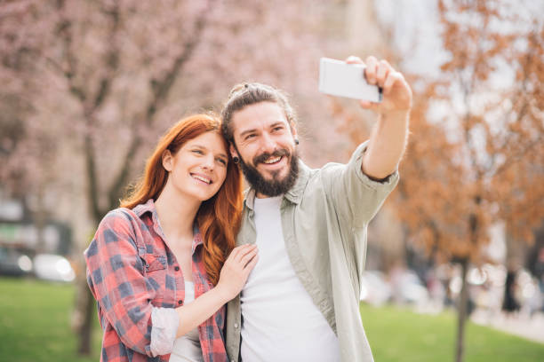 istock Young couple looking at mobile phone while taking selfie at the park 1005355734