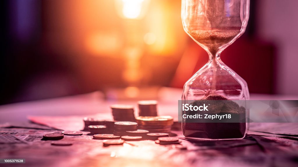 Sand running through the shape of hourglass on table with banknotes and coins of international currency. Time investment and retirement saving. Urgency countdown timer for business deadline concept Retirement Stock Photo