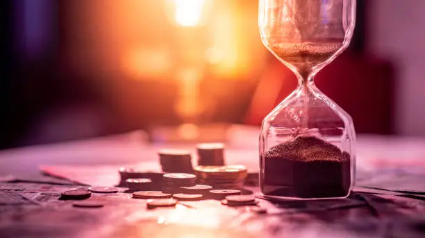 Photo of Sand running through the shape of hourglass on table with banknotes and coins of international currency. Time investment and retirement saving. Urgency countdown timer for business deadline concept