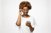 Closeup photo of African American woman standing isolated on gray background looking at screen of cellphone, browsing web pages and smiling nicely while chatting