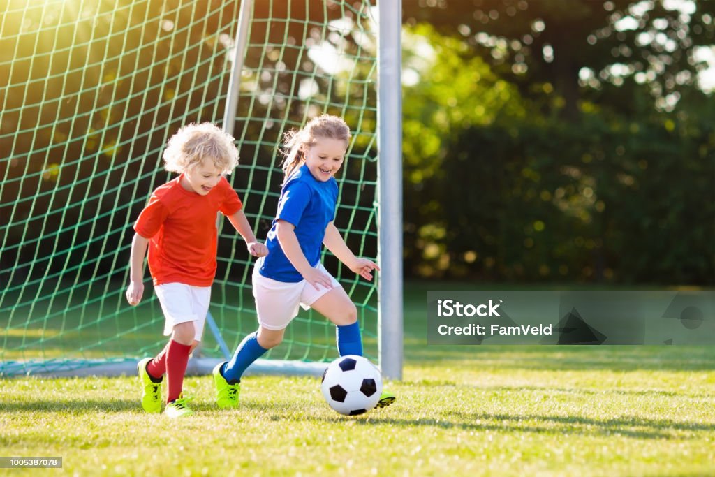 Kids play football. Child at soccer field. Kids play football on outdoor field. Children score a goal at soccer game. Girl and boy kicking ball. Running child in team jersey and cleats. School football club. Sports training for young player. Child Stock Photo
