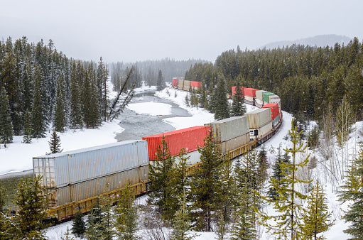 Train carrying Containers alongside a Frozen Mountain River on a Snowy Winter Day. Banff National Park, AB, Canada.