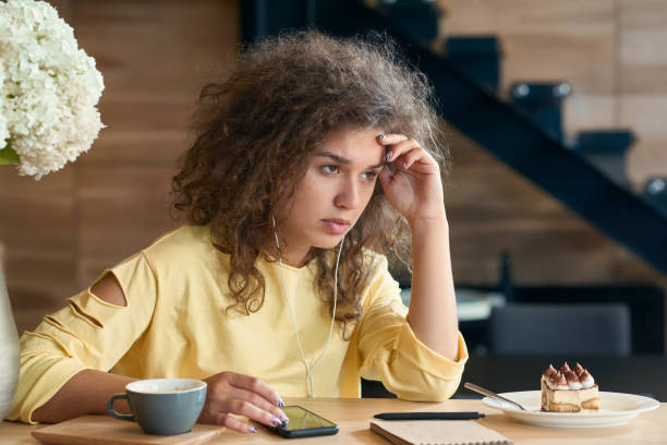 Depressed young woman with curly hair sitting on wooden restaurant's table. Depressed young woman with curly hair sitting on wooden restaurant's table, looking exhausted and angry, feeling negative emotions. Cup of coffee and white lilac flowers, stairs on background. narcolepsy stock pictures, royalty-free photos & images