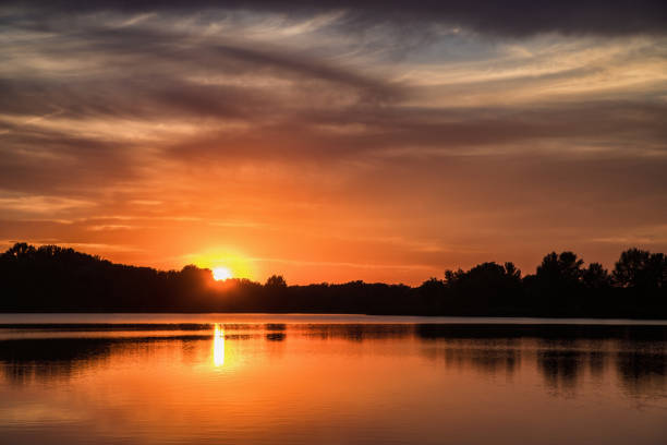 Setting sun and sunset clouds over silhouettes of trees with reflection in water surface of Sulianske jazero lake, Slovakia.