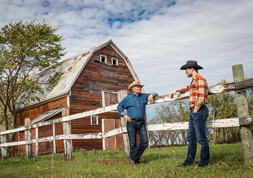 two farming caucasian cowboys standing by a wooden fence having a discussion in front of a red barn on a warm bright sunny day in summer.