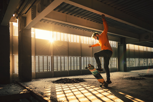 Extreme street sport. Hipster man jumping and riding on skateboard at an abandoned building at sunset. Image with grain