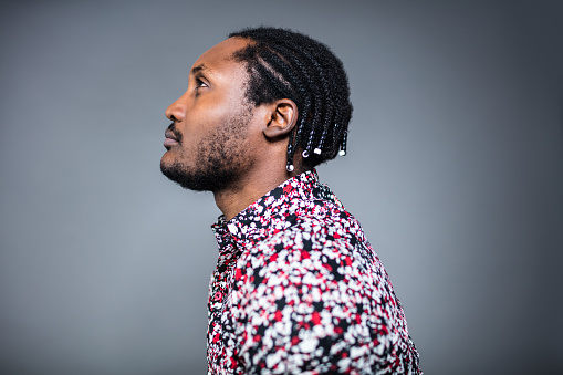 Side view of confident mid adult man with braided black hair. Male is looking away against gray background. He is in red pattern shirt.