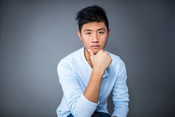 Portrait of confident young man sitting against gray background. Handsome male is with hand on chin. He is in blue t-shirt.