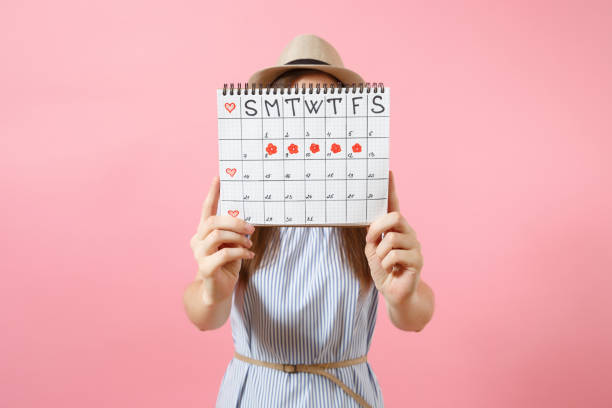 retrato de mujer con vestido azul cubre cara, escondiéndose detrás de calendario de periodos para comprobar los días de menstruación aisladas sobre fondo rosa tendencias. concepto médico, atención médico, ginecológico. copia espacio. - menstruación fotografías e imágenes de stock