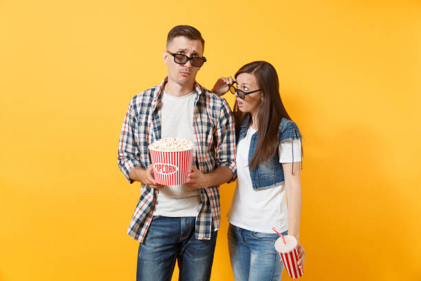 młoda wyczerpana para kobieta i mężczyzna w okularach 3d oglądania filmu na randce gospodarstwa wiadro popcorn i plastikowy kubek sody lub coli izolowane na żółtym tle. emocje w kinie. - drink bottle soda bucket zdjęcia i obrazy z banku zdjęć