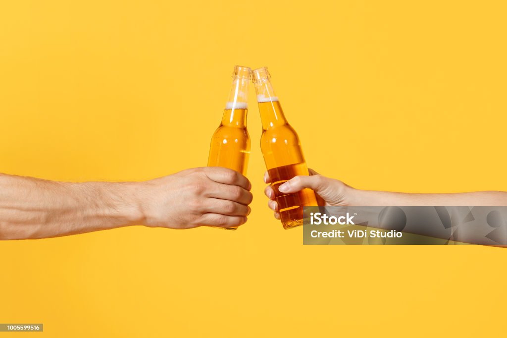 Close up cropped of woman and man two hands horizontal holding lager beer glass bottles and clinking isolated on yellow background. Sport fans cheer up. Friends leisure lifestyle concept. Copy space. Close up cropped of woman and man two hands horizontal holding lager beer glass bottles and clinking isolated on yellow background. Sport fans cheer up. Friends leisure lifestyle concept. Copy space Beer - Alcohol Stock Photo