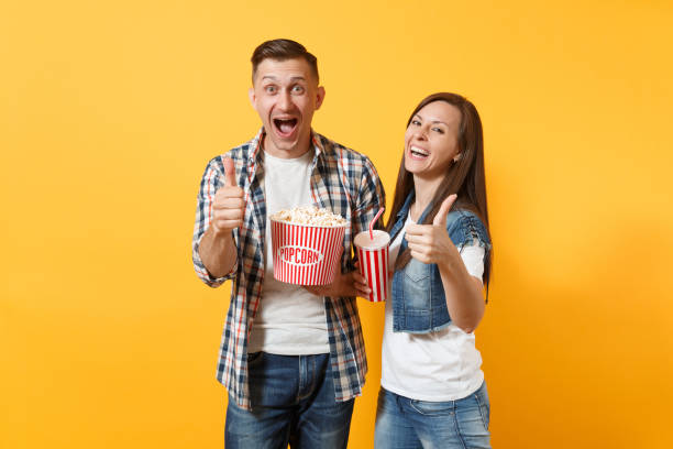 młoda uśmiechnięta para kobieta kobieta oglądania filmu na dzień gospodarstwa wiadro popcorn plastikowy kubek sody lub cola pokazano kciuki w górę izolowane na żółtym tle. emocje w kinie. - drink bottle soda bucket zdjęcia i obrazy z banku zdjęć