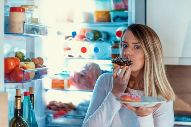 Photo of a young woman doing midnight snack at home. She eats a donuts and looks for food into the refrigerator at night. Close up portrait of a hungry greedy girl eating donuts in front of open fridge. Hungry attractive girl eating donuts at night near fridge
