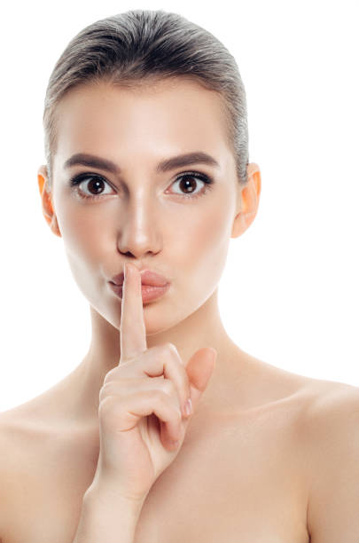 Cropped image of a young woman holding index finger at her lips, saying 'shh' on a white studio background. side profile view