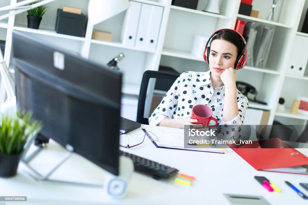 Una joven se sienta en auriculares en una mesa en la oficina, tiene una taza roja en sus manos y mira el monitor. - Foto de stock de Largometrajes libre de derechos