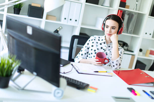 une jeune fille se trouve dans le casque à une table dans le bureau, tient une tasse rouge dans ses mains et se penche sur le moniteur. - the thinker audio photos et images de collection