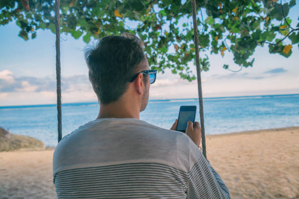 istock Man sitting on the wooden swing and using cellphone on the tropical beach. 1005806064