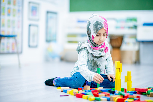 A Middle-eastern girl is sitting on the floor in her elementary school classroom. She is playing with blocks, and smiling at the camera while building towers.