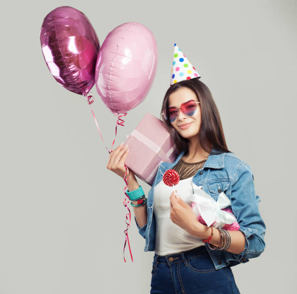 cheerful woman with gift box, candy lollipop, birthday cap and balloons. young happy female model on holiday background with copy space - personal accessory balloon beauty birthday imagens e fotografias de stock