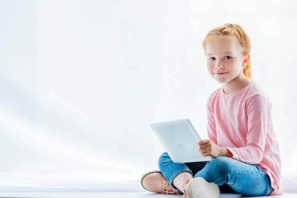 istock beautiful redhead child using digital tablet and smiling at camera while sitting on windowsill 1005935152