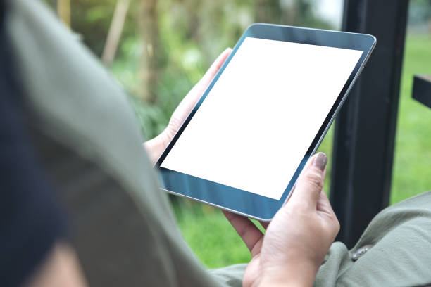 istock Mockup image of woman's hands holding black tablet pc with white blank screen while sitting in cafe with green nature background 1005941824