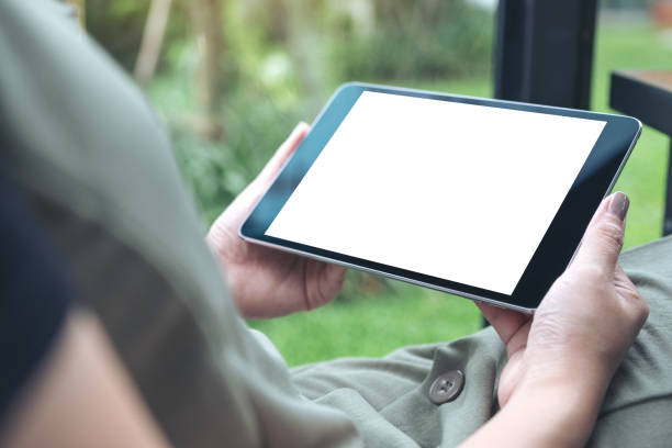 istock Mockup image of woman's hands holding black tablet pc with white blank screen while sitting in cafe with green nature background 1005941828