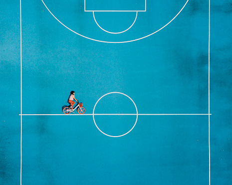 Cycling in a surreal world. Aerial view. Conceptual realism trend. Young adult woman cycling in a basketball court.