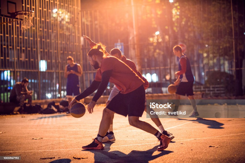 They know to play streetball Young men playing basketball on courtyard at dusk. They  play basketball on courtyard, performing exhibitions and slam dunking Basketball - Sport Stock Photo