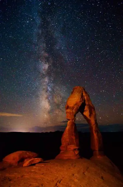Summer nights in Arches national park, with the Milky Way rising to be perpendicular with Delicate Arch (lit by a separate photo tour). The b/g glow is Moab, 8 miles away.
