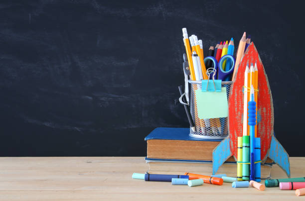 istock Back to school concept. cardboard rocket and pencils over open book in front of classroom blackboard. 1006058532