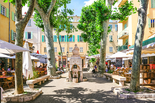 Menton, France - May 31, 2018: People on little square with bars, cafés and restaurants in the shade of trees and small statue in the center of a town on French Riviera, Cote d'Azur.