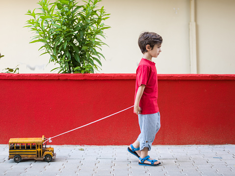 5 Years old little boy wearing a red t-shirt and blue pants pulling a toy yellow school bus on sidewalk in outdoor. The background is red colored wall. Shot in outdoor daylight with a medium format camera.