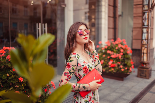 Young woman wearing classic dress with accessories outdoors. Beauty fashion model in glasses holding handbag by hotel entrance