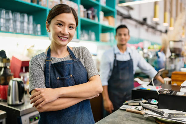 Proud Smiling Woman Coffee Shop Owner Happy and confident smiling chinese woman, proud small business coffee shop owner standing with arms crossed behind the counter. Young man, the barsita - coffee maker - standing in the background of the stylish cafe in Kuala Lumpur, Malaysia chinese restaurant stock pictures, royalty-free photos & images