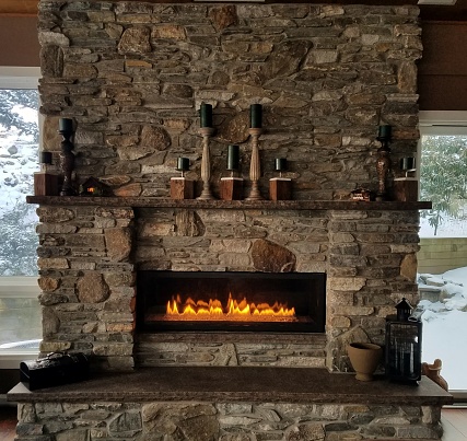 A rustic setting of a large stone fireplace with mantle and hearth and a napoleon landscape style fireplace lit with burning flames; Winter, cozy, rustic, design, interiors, stone, masonry ideas.