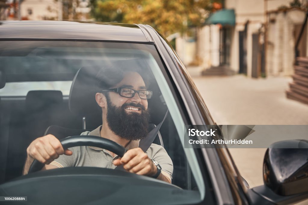 A modern bearded man driving a car A Young attractive man driving a vehicle, looking at scenery, seen through the windshield glass. Happy driver, holding hands on the steering wheel with a wide beautiful smile. Car Stock Photo