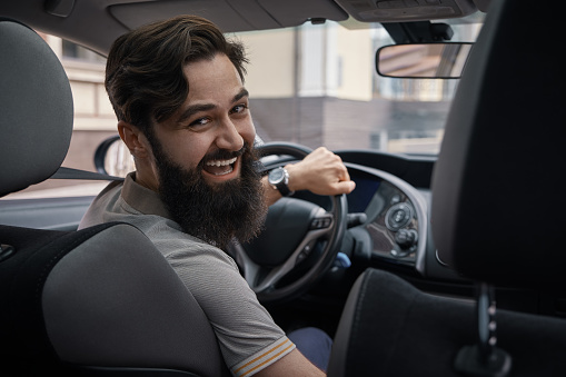 Riding his new car. Side view of handsome young bearded man driving his car and smiling, in the city