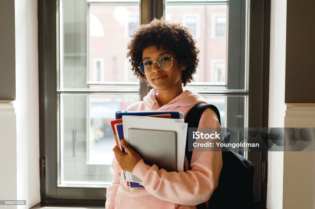 Beautiful young woman with books and digital tablet standing at window in campus Student Stock Photo