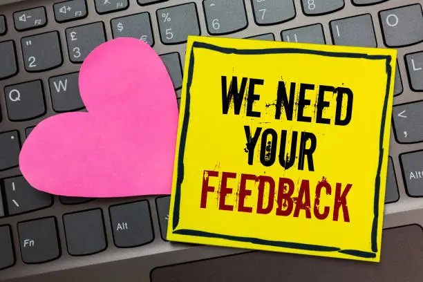 Handwriting text writing We Need Your Feedback. Concept meaning Give us your review thoughts comments what to improve Written on black bordered yellow page touched pink heart on computer keyboard
