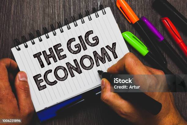 Word Writing Text The Gig Economy Business Concept For Market Of Shortterm Contracts Freelance Work Temporary Hand Holding Pen And Paper Sketch Words Near Lie Some Pen On Woody Desk Stock Photo - Download Image Now