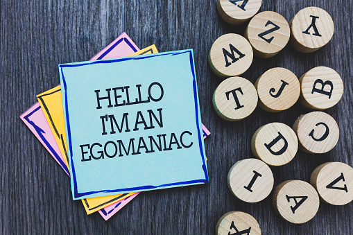 Handwriting text Hello I am An Egomaniac. Concept meaning Selfish Egocentric Narcissist Self-centered Ego Black wooden deck written sticky note beside some round woody alphabets