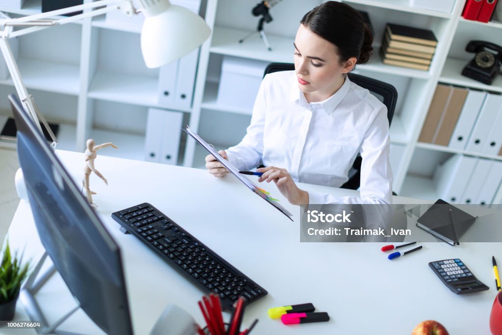 Une jeune fille dans le Bureau tient un stylo dans sa main et travaille avec des documents et un ordinateur. - Photo de Affaires libre de droits