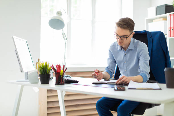 istock A young man sits in the office at a computer desk and writes in a notebook. 1006276652