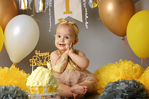 Portrait of cute adorable Caucasian baby girl in tutu tulle skirt celebrating her first birthday. Cake smash concept. Child sitting on floor in studio, balloons, eating dessert