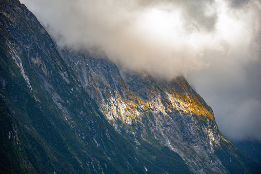 Misty and cloudy sky covering mountain top with some sunlight shining through at Milford Sounds, New Zealand.